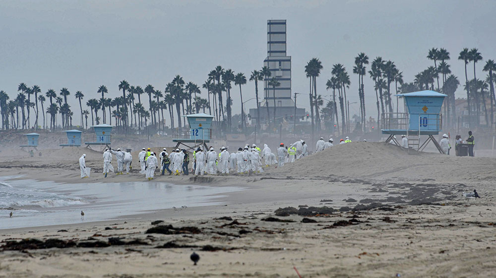 Oil spill workers in white protective gear clean oil from a sandy beach. 
