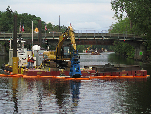 A barge with a earth mover scoops sediment from a river with a bridge in background. 