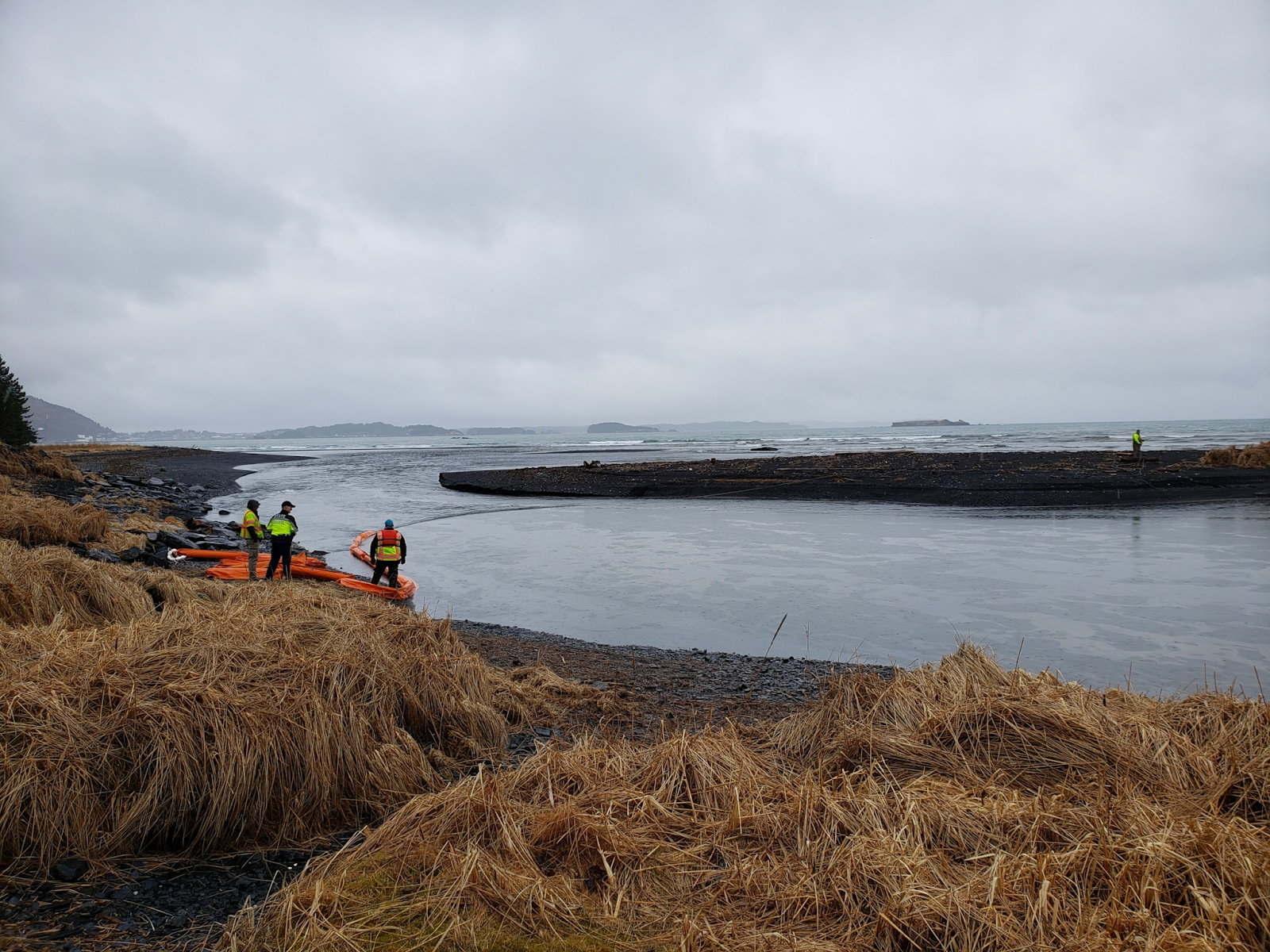 Three people stand on the edge of the water with oil spill boons deployed. 