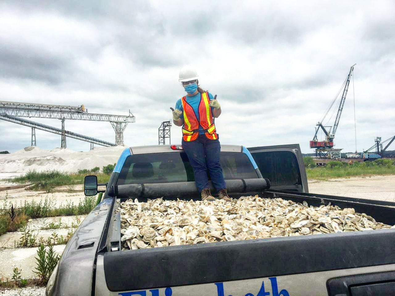 A woman in a mask and gloves gives a thumbs up while standing on a truck pickup-bed full of white oyster shells. 