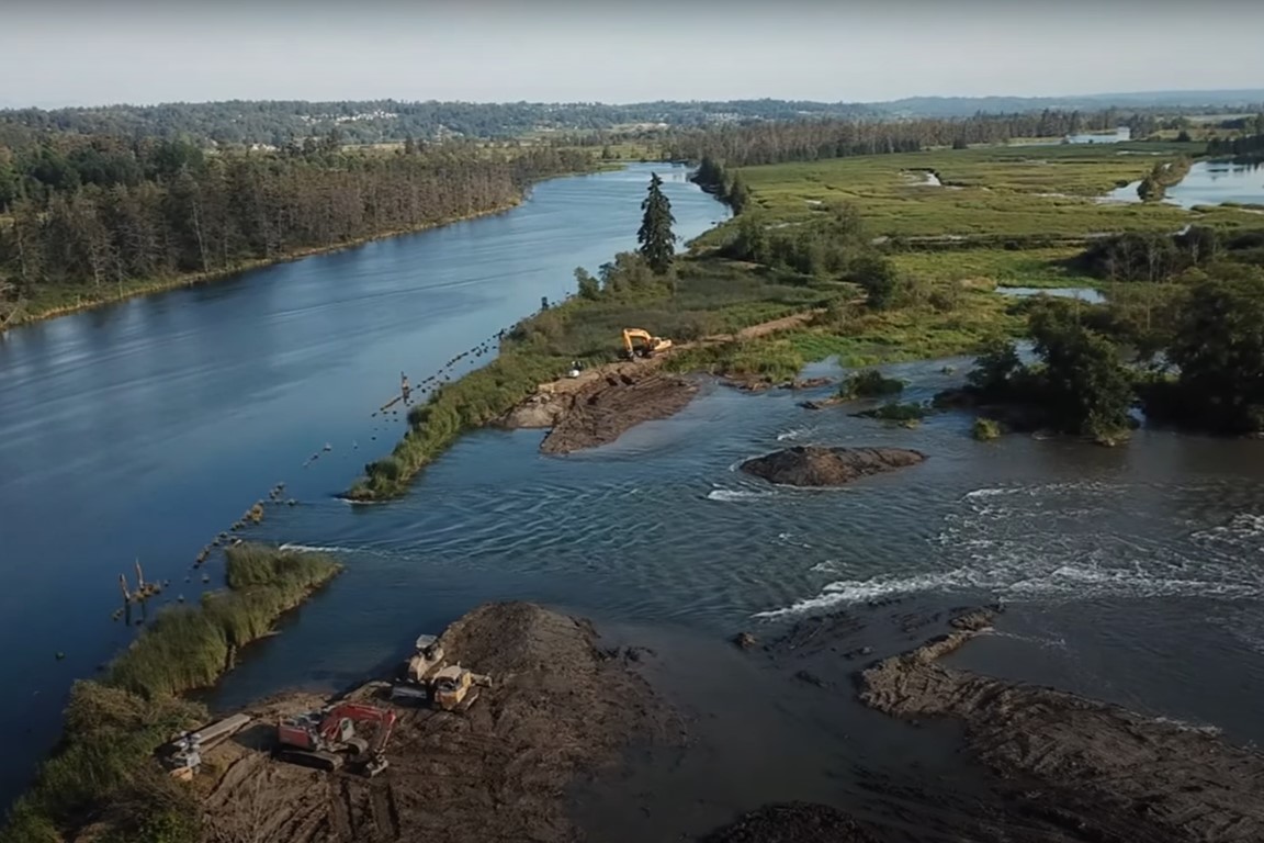 This image is taken from an airborne drone. You can see upstream on the left. In the foreground you can see the breach in the levee where river water is pouring into the site. Heavy equipment is parked on either side of the breach.