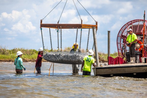 a crew lowers a large basket of rocks into the water 