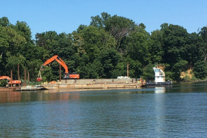 Two excavator machines work on building a shoreline project. A barge holding materials is in the water offshore.