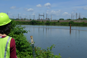 Woman in a vest and hard hat stands at the edge of a river with industrial site in background. 