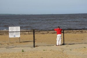 Laurence Harbor, New Jersey waterfront with an EPA public health hazard sign and a person in a red shirt and white pants standing behind a fence that restricts access to the beach.