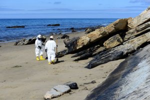Two cleanup workers in white protective gear and yellow boots stand next to oiled rocks and ocean in background. 